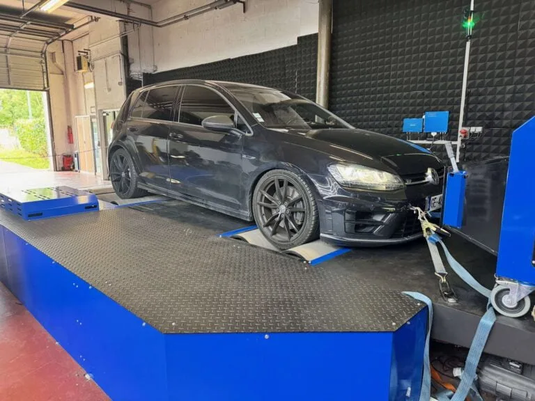 Une Volkswagen Golf VIII R 2.0 TSI dans un centre de reprogrammation moteur. La voiture est attachée par des sangles pour la sécurité pendant le test. L'environnement de l'atelier inclut des murs soufflés avec des dispositifs de mesure et un ventilateur visible à l'arrière-plan pour refroidir le véhicule pendant le test.